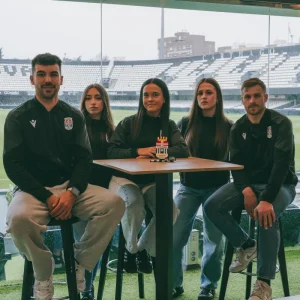 Foto grupal de cinco jugadores y jugadoras del FC Cartagena sentados alrededor de una mesa alta, con un modelo de LEGO de la torre del estadio del FC Cartagena en el centro, posando para la cámara con el estadio de fondo.