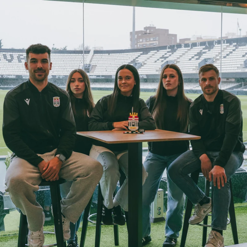 Foto grupal de cinco jugadores y jugadoras del FC Cartagena sentados alrededor de una mesa alta, con un modelo de LEGO de la torre del estadio del FC Cartagena en el centro, posando para la cámara con el estadio de fondo.