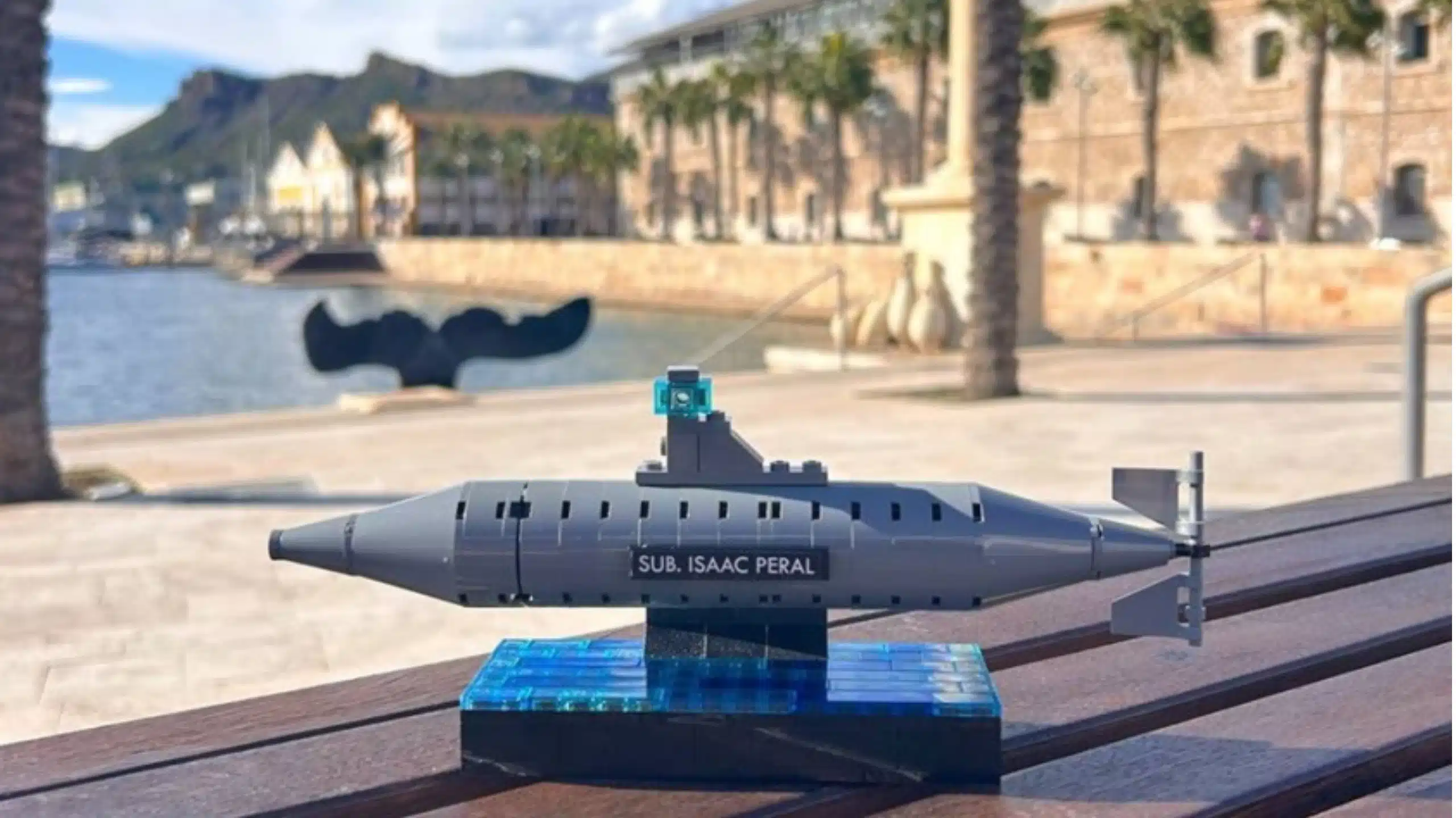 Set con bloques LEGO® del submarino Isaac Peral sobre una base azul, colocada en primer plano sobre una mesa de madera en el puerto de Cartagena, con el paseo marítimo, palmeras, edificios históricos y montañas desenfocados al fondo.